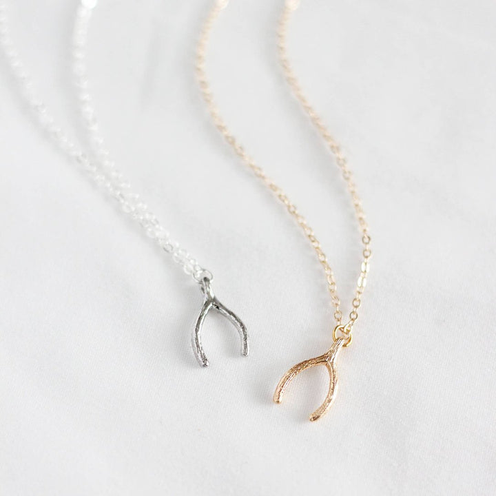 Two necklaces on a white surface. One necklace has a gold wishbone pendant, and the other has a silver wishbone pendant. Both necklaces have delicate chains.