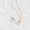 Two necklaces on a white surface. One necklace has a gold wishbone pendant, and the other has a silver wishbone pendant. Both necklaces have delicate chains.