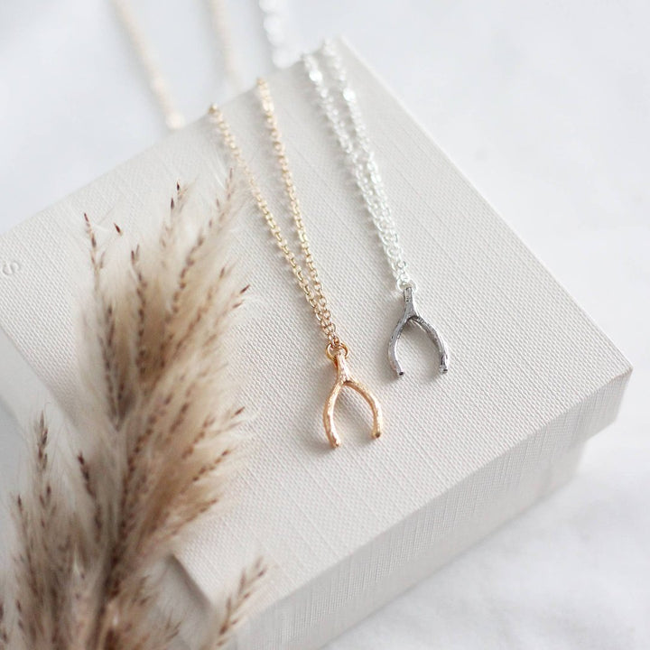 Two necklaces on a white surface. One necklace has a gold wishbone pendant, and the other has a silver wishbone pendant. Both necklaces have delicate chains.
