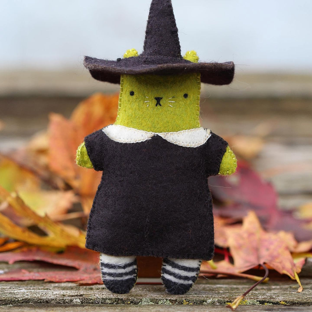 A small felt witch cat figurine with a dark brown hat and dress, olive green face and hands, and striped socks, standing on a rustic wooden surface with autumn leaves in the background.