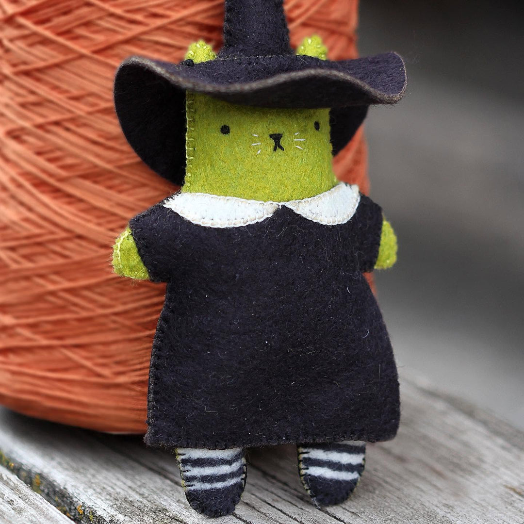 A handmade felt cat doll wearing a black witch hat and dress with a white collar, standing in front of a large orange yarn ball on a weathered wooden surface.