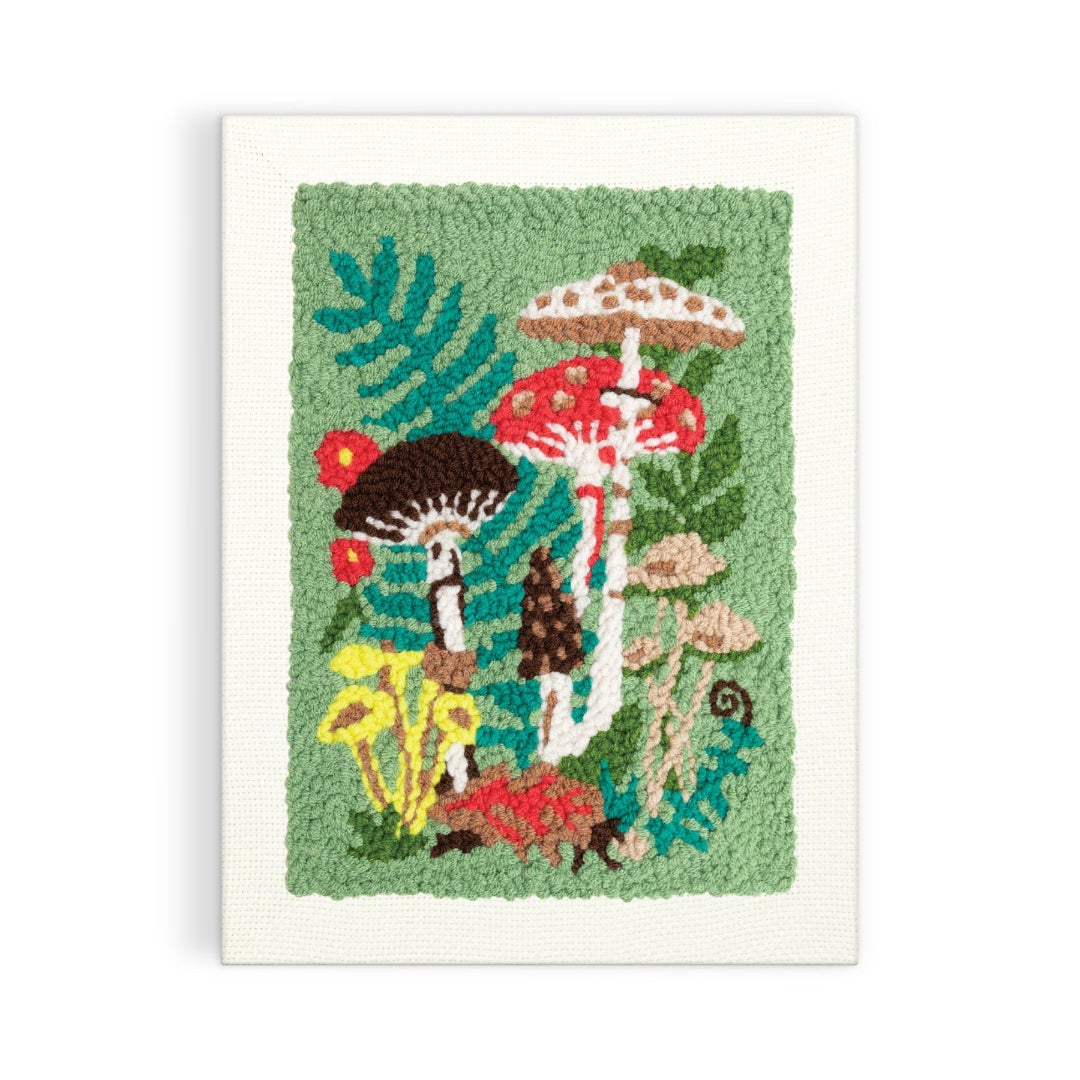 A square wall-hanging or rug-style textile artwork featuring colorful mushrooms and green foliage on a textured light green field, bordered by a white stitched edge, shown against a plain white background.