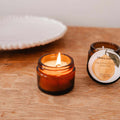 Candle in a glass jar on a wooden surface with another candle and a dish in the background.