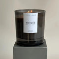 A black glass jar candle with a white label. The label reads "NORTH + ROSE" and "WREATH" with "Balsam - Pine - Raspberry" underneath. The candle is sitting on a black box with the same label details.