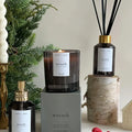 A black glass jar candle with a white label. The label reads "NORTH + ROSE" and "WREATH" with "Balsam - Pine - Raspberry" underneath. The candle is sitting on a black box with the same label details.