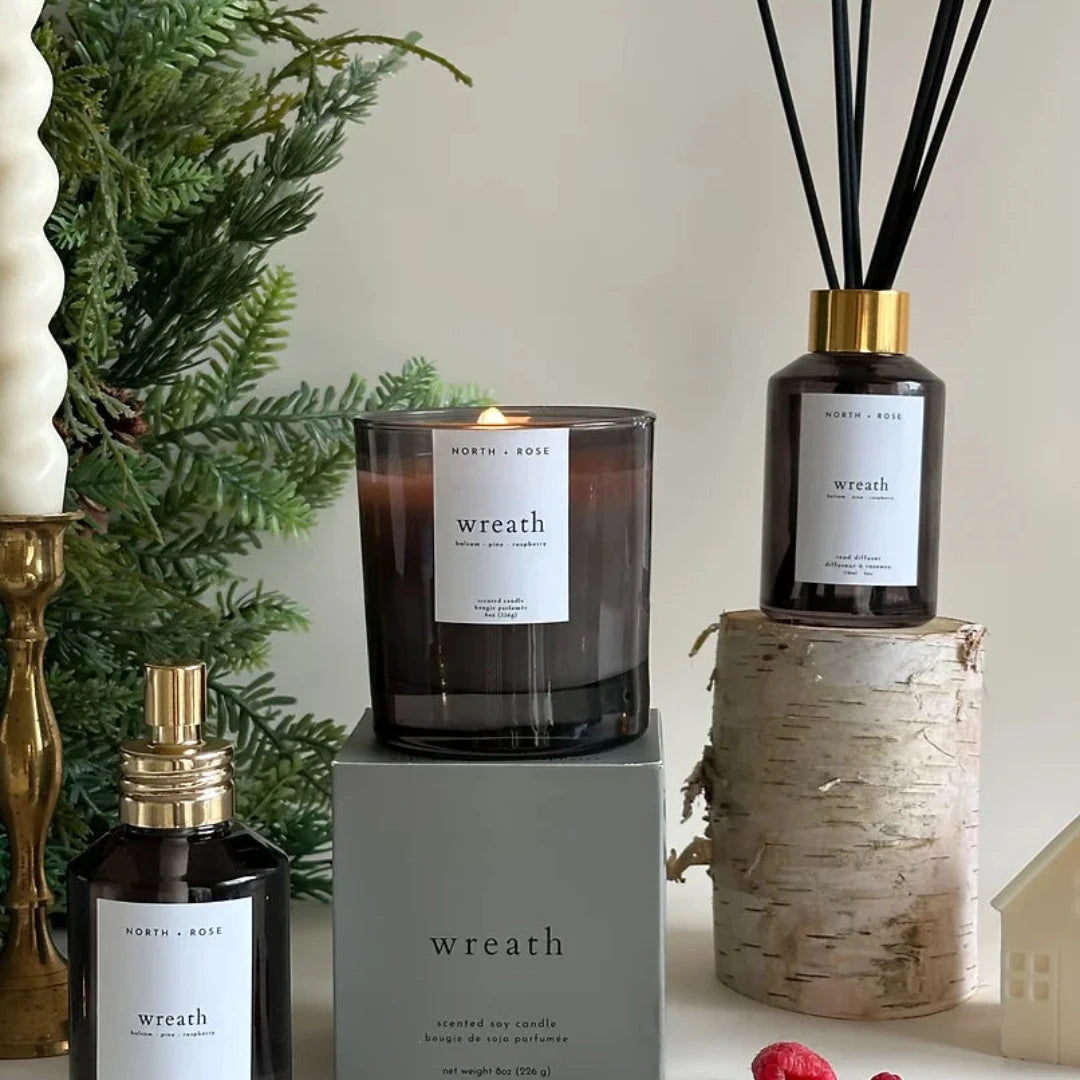 A black glass jar candle with a white label. The label reads "NORTH + ROSE" and "WREATH" with "Balsam - Pine - Raspberry" underneath. The candle is sitting on a black box with the same label details.