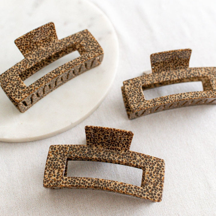 Three large hair claw clips in a leopard print pattern on a white marble surface.