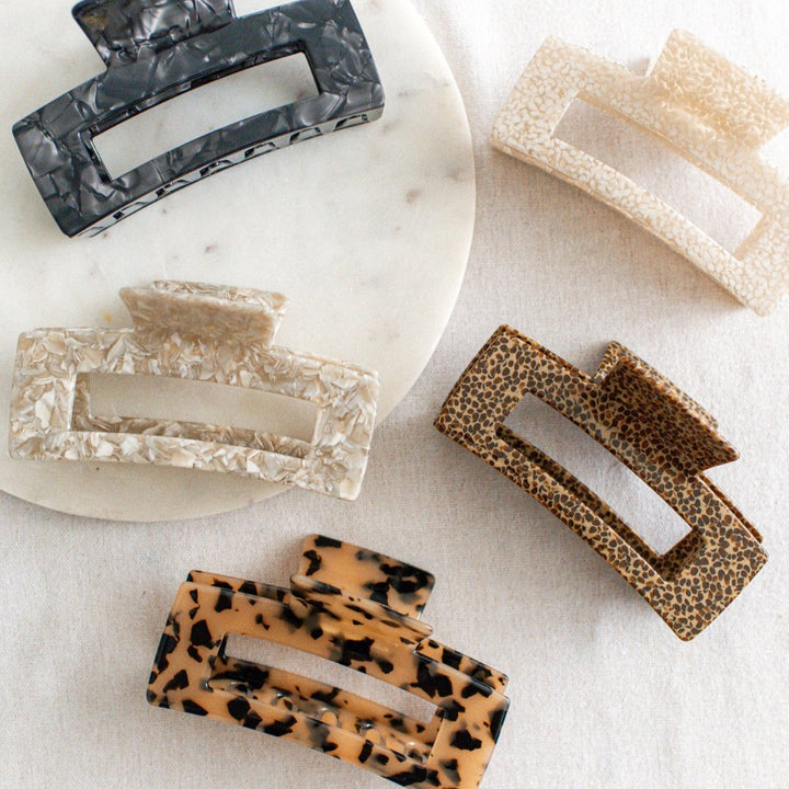Five hair claw clips on a white marble surface. The clips are in various colors and patterns, including black marbled, beige marbled, brown and black leopard print, and a light brown marbled pattern.