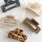 Five hair claw clips on a white marble surface. The clips are in various colors and patterns, including black marbled, beige marbled, brown and black leopard print, and a light brown marbled pattern.