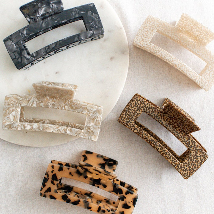 Four hair claw clips on a white marble surface. The clips are in various colors and patterns, including black marbled, beige marbled, brown and black leopard print, and a light brown marbled pattern.