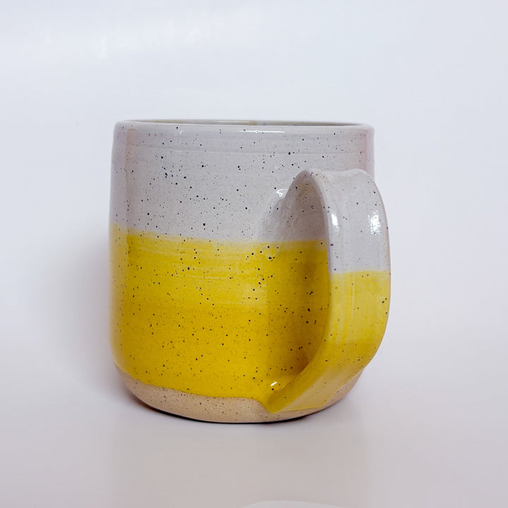Yellow Dipped Mug by Valerie Daoust, a handmade ceramic mug with a colorblock design featuring a light gray top and a speckled yellow middle section, displayed on a light gray background. The mug has a large, rounded handle, a speckled texture, and is made in Mississippi Mills, Ontario, Canada.