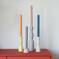 Four colorful striped taper candles displayed in decorative sculptural holders on a red wooden cabinet against a light gray wall.