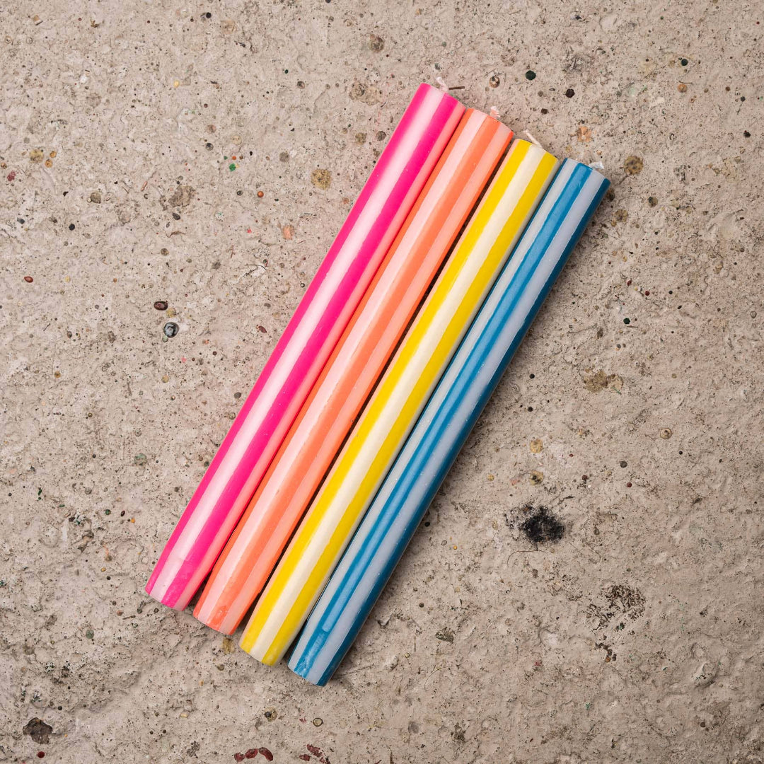 Five striped candles in pink, orange, yellow, and blue, arranged diagonally on a textured concrete surface.