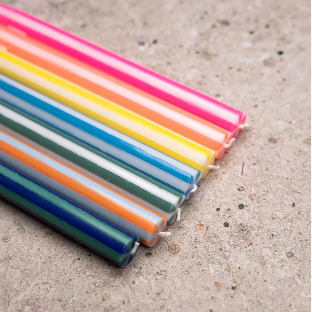 A set of colorful taper candles arranged diagonally on a textured concrete surface. The candles feature multiple vibrant colors including pink, yellow, orange, green, blue, and white.