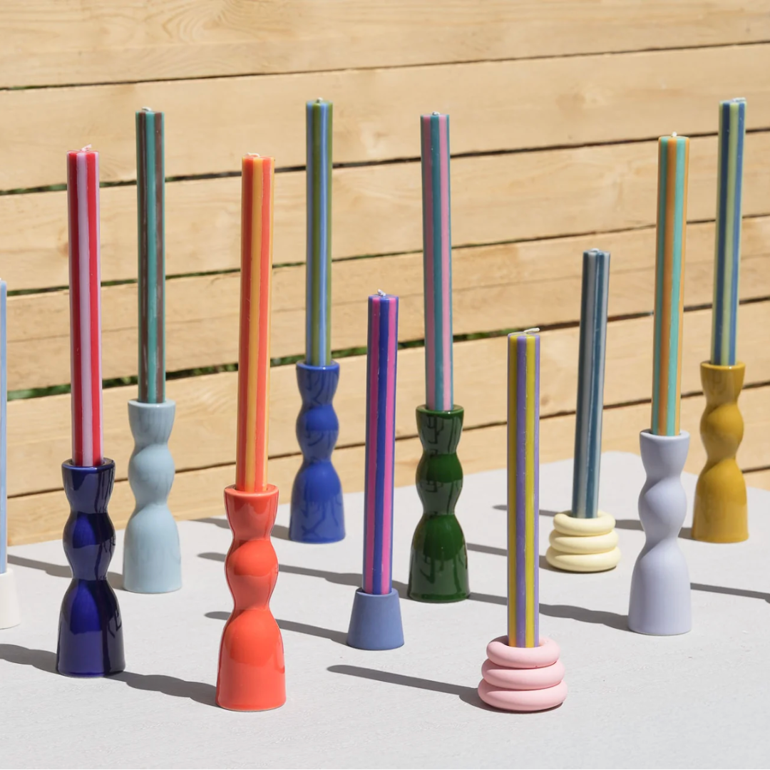 A set of colorful, sculptural candlesticks with twisted handles and long cylindrical stems, displayed on a white surface against a light wood slat wall.