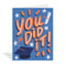 Blue greeting card featuring colourful illustrations by Erin Mac of a graduation cap with an orange tassel, a rolled diploma tied with an orange ribbon, orange bursts of light, and white stars surrounding the large, stylized orange and white text 