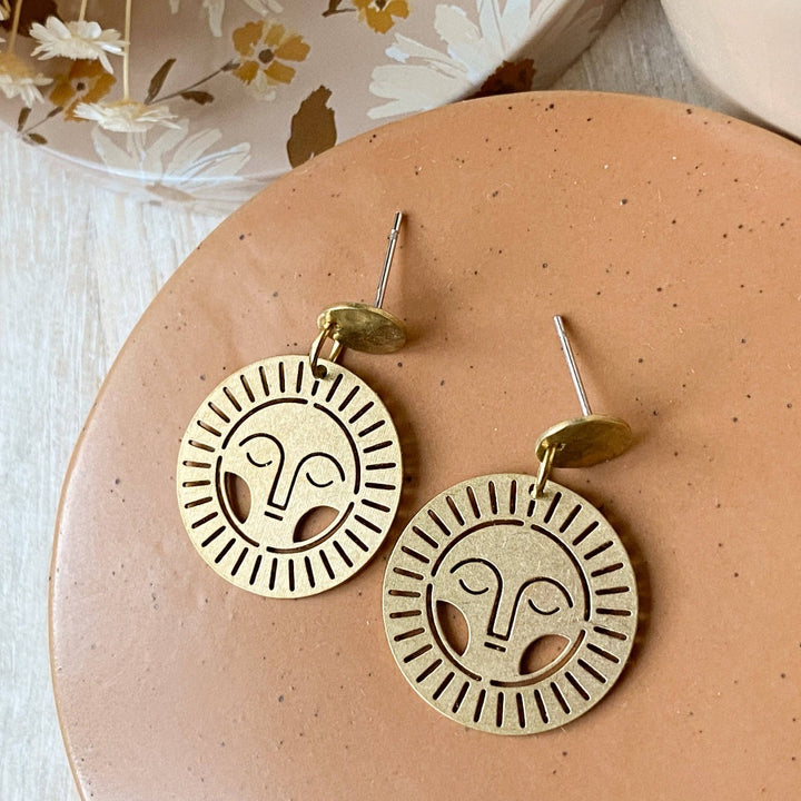 Brass Viviette Earrings. These elegant earrings feature circular brass discs with a sunburst cutout design and a small hammered brass circle at the top. The earrings are displayed on a soft, neutral-toned surface, adding a warm and sophisticated touch.