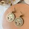 Brass Viviette Earrings. These elegant earrings feature circular brass discs with a sunburst cutout design and a small hammered brass circle at the top. The earrings are displayed on a soft, neutral-toned surface, adding a warm and sophisticated touch.
