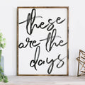 Framed wood sign with the text "These are the days" in black brush script against a white background.