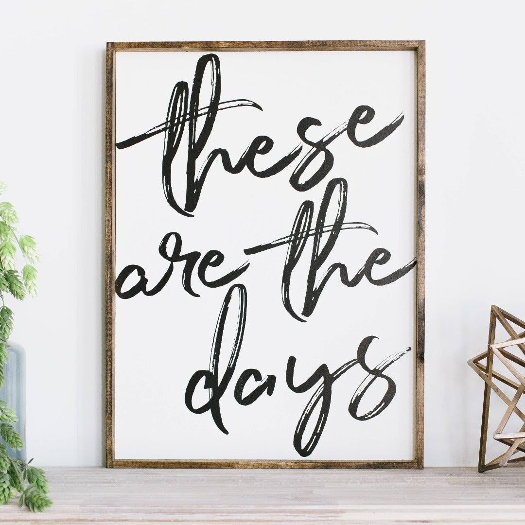 Framed wood sign with the text "These are the days" in black brush script against a white background.