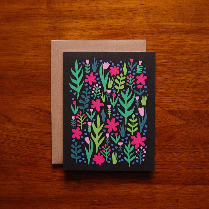 Bright and cheerful greeting card featuring a lively pattern of various pink, green, and blue flowers and leaves against a black background. The card is accompanied by a kraft paper envelope in the background, adding a rustic and natural touch.