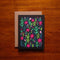 Bright and cheerful greeting card featuring a lively pattern of various pink, green, and blue flowers and leaves against a black background. The card is accompanied by a kraft paper envelope in the background, adding a rustic and natural touch.