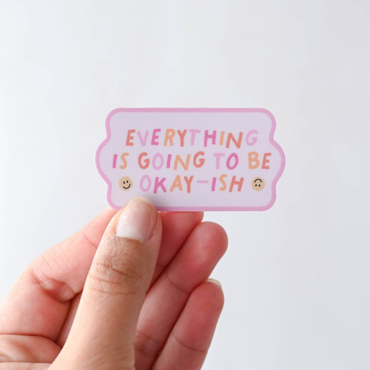 A hand holding an Everything Is Going To Be Okay-ish Sticker, a pink sticker with colourful words reading 