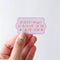 A hand holding an Everything Is Going To Be Okay-ish Sticker, a pink sticker with colourful words reading 