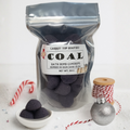 Bag of 'Coal' bath bomb gumdrops with a label, candy cane, and ornament on a white background