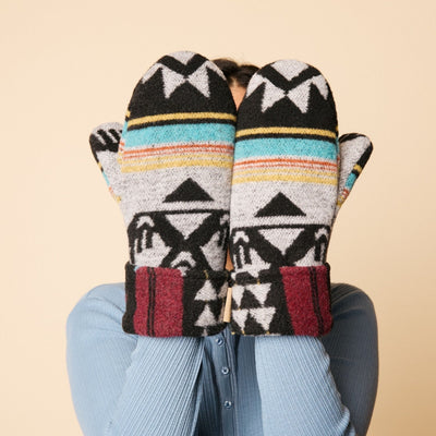 Pair of patterned mittens held by a person wearing a blue sweater against a beige background