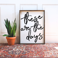 Framed wood sign with the text "These are the days" in black brush script against a white background.