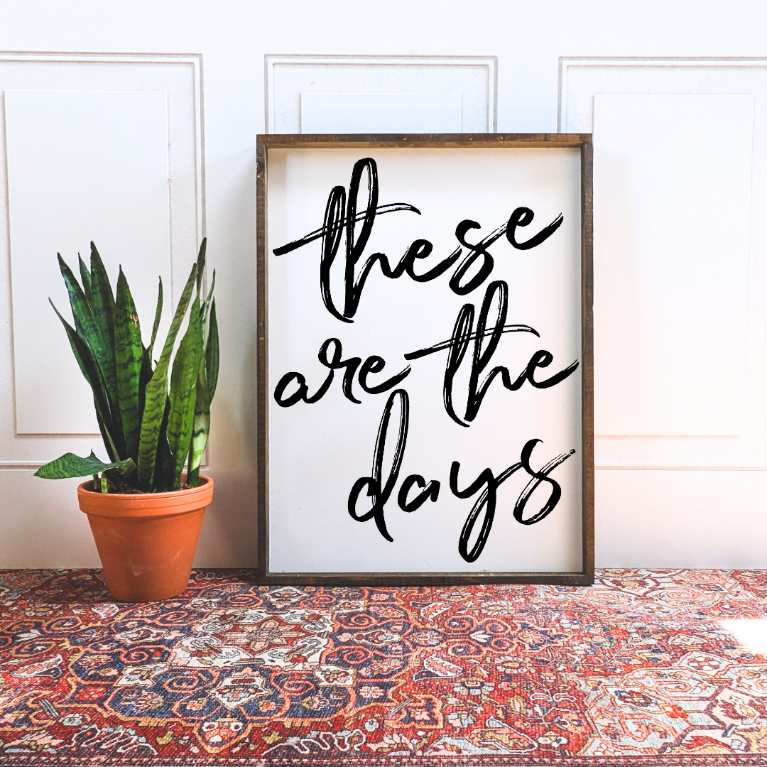 Framed wood sign with the text "These are the days" in black brush script against a white background.