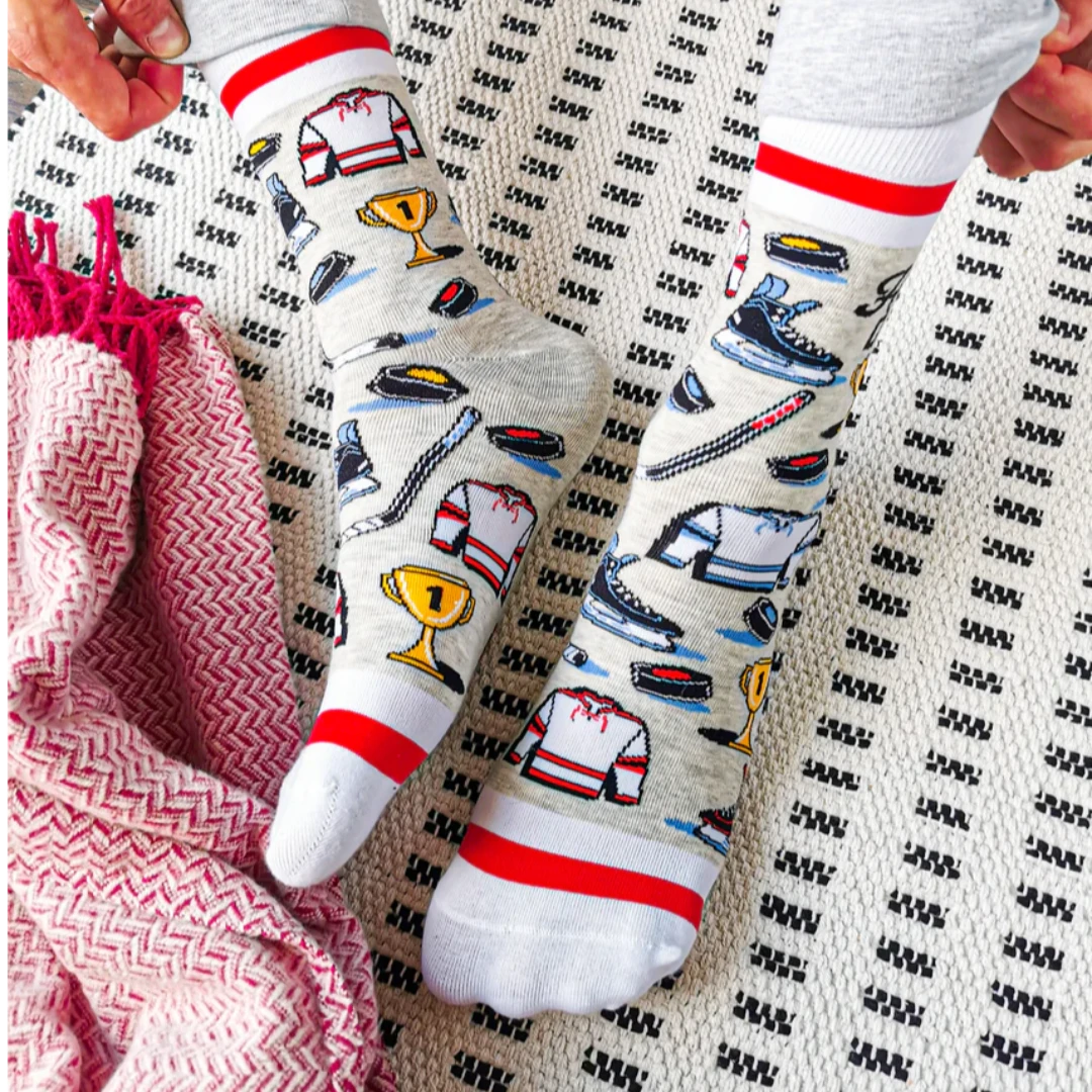 A pair of crew socks featuring a hockey theme. The socks are mostly grey with white and red stripes at the top and bottom. The design includes hockey-related icons like a trophy, skates, and a puck. The text "Hockey Dad" is printed on the socks.