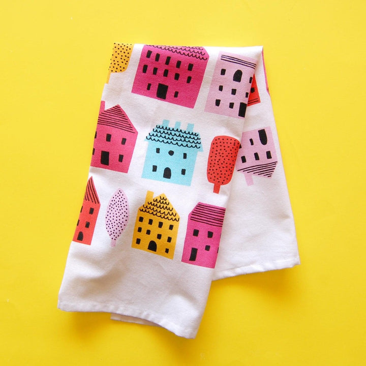 A white tea towel with a pattern of colorful houses and trees. The houses are in various sizes and colors, including pink, red, orange, yellow, blue, and green. The trees are green and have simple leaf shapes.