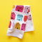 A white tea towel with a pattern of colorful houses and trees. The houses are in various sizes and colors, including pink, red, orange, yellow, blue, and green. The trees are green and have simple leaf shapes.