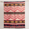 A Kwe Indigenous reversible blanket featuring bold red, black, white, and ochre geometric patterns, hanging from a natural branch. This culturally significant blanket is available in Ottawa, Ontario, Canada.