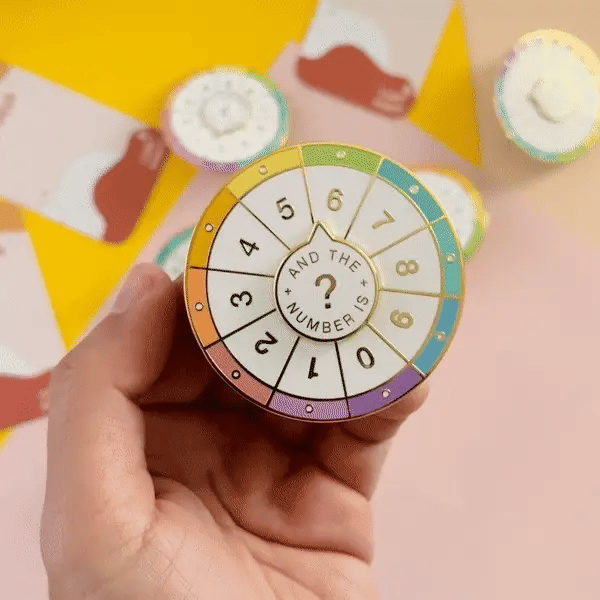 A circular pin with a colorful wheel featuring numbers from 0 to 9. The phrase 