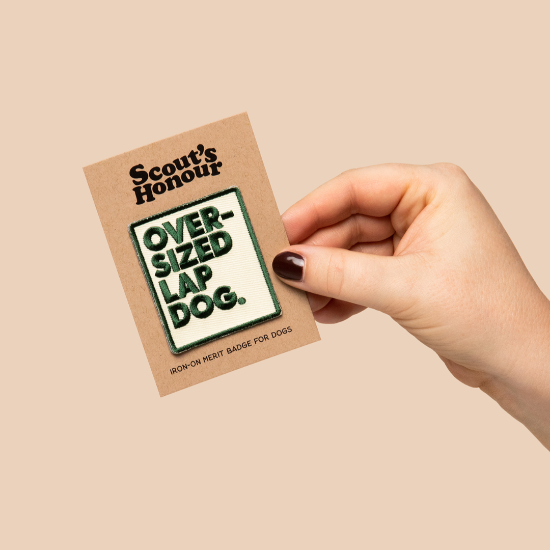 Hand holding a 'Scout's Honour' over-sized lap dog badge against a beige background