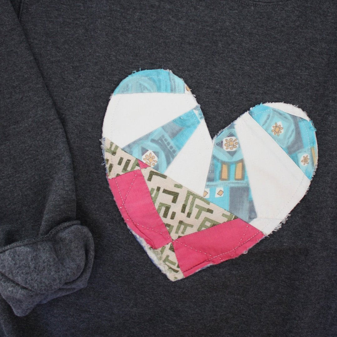 a grey sweatshirt with a large, heart-shaped patch. The patch is made from various fabrics with various patterns, sewn together in a patchwork style.