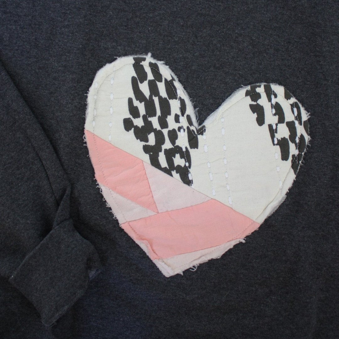 a grey sweatshirt with a large, heart-shaped patch. The patch is made from various fabrics with various patterns, sewn together in a patchwork style.