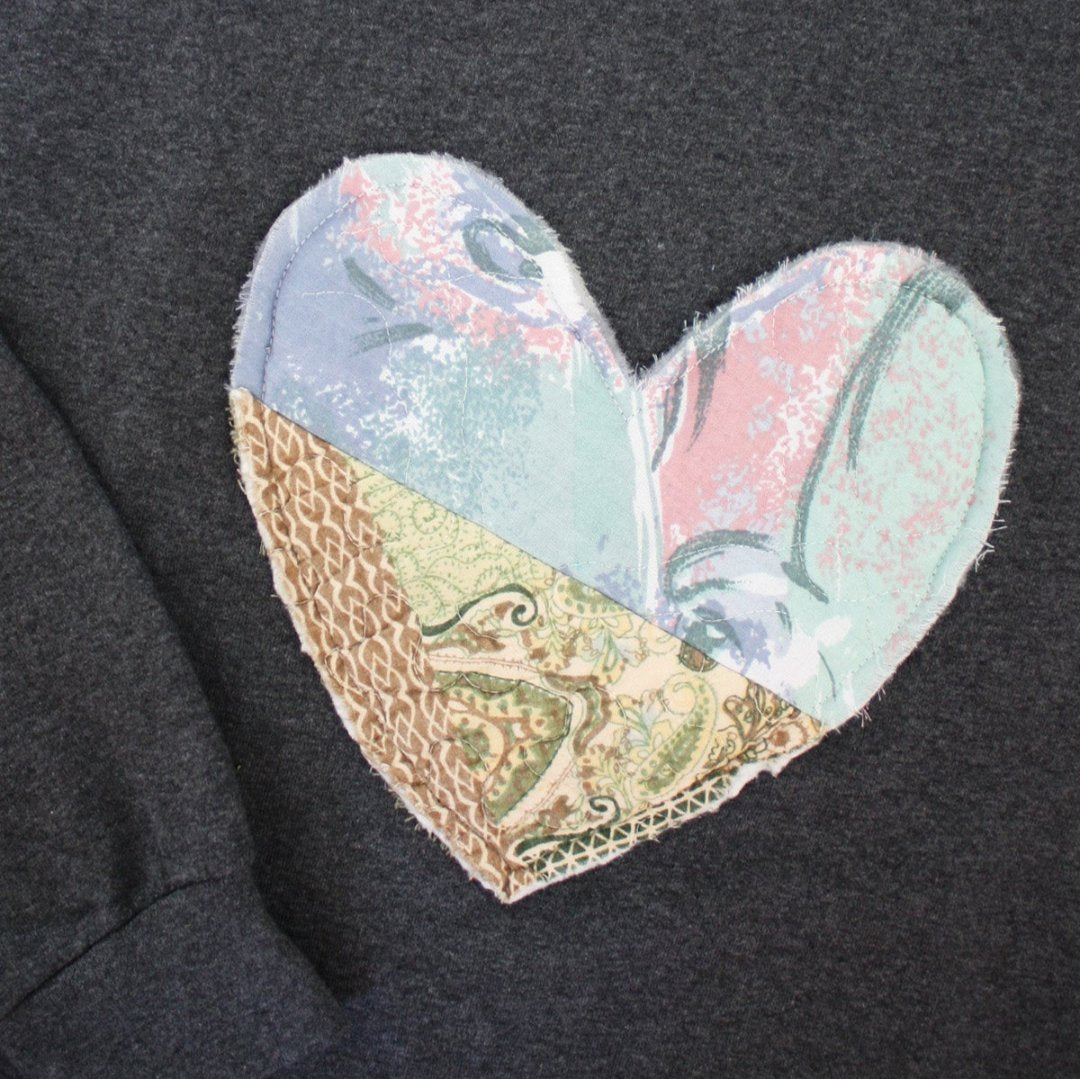 a grey sweatshirt with a large, heart-shaped patch. The patch is made from various fabrics with various patterns, sewn together in a patchwork style.