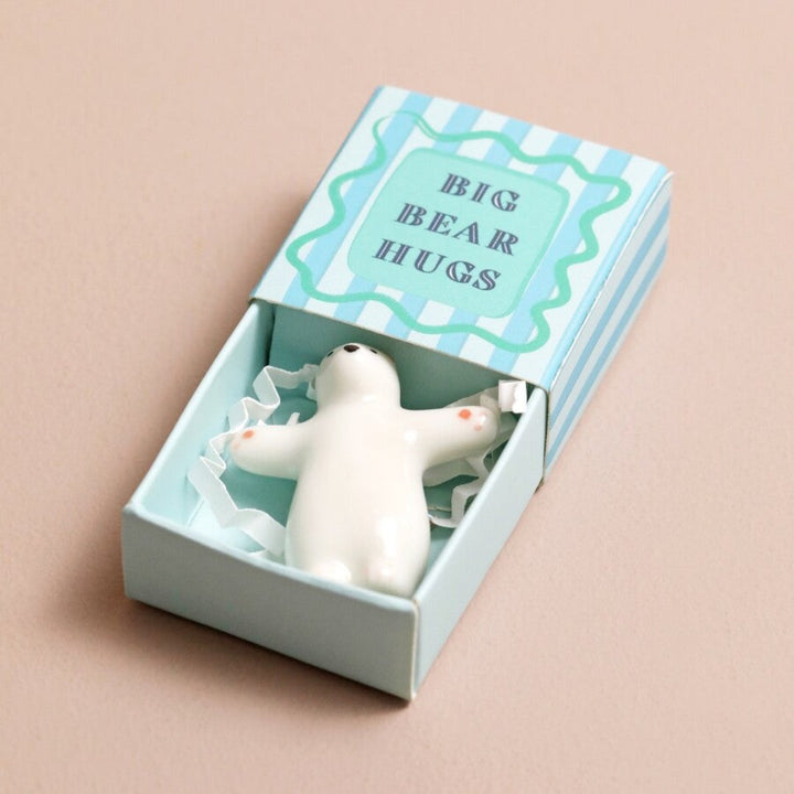 A small, white ceramic bear figurine inside a blue matchbox. The bear is in a hugging pose, and the box has the text 