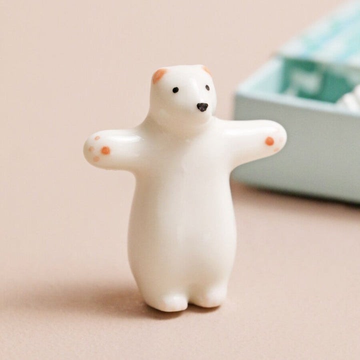 A small, white ceramic bear figurine inside a blue matchbox. The bear is in a hugging pose, and the box has the text 