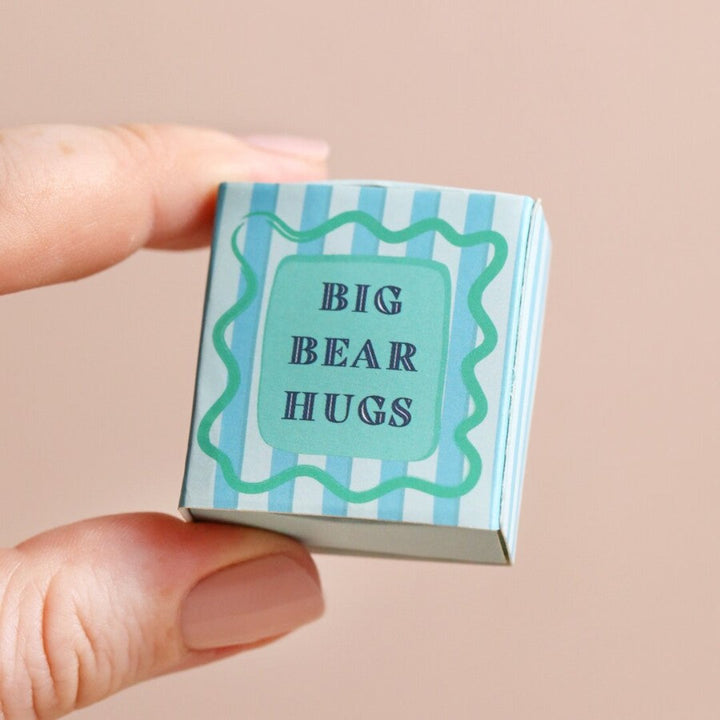 A small, white ceramic bear figurine inside a blue matchbox. The bear is in a hugging pose, and the box has the text 