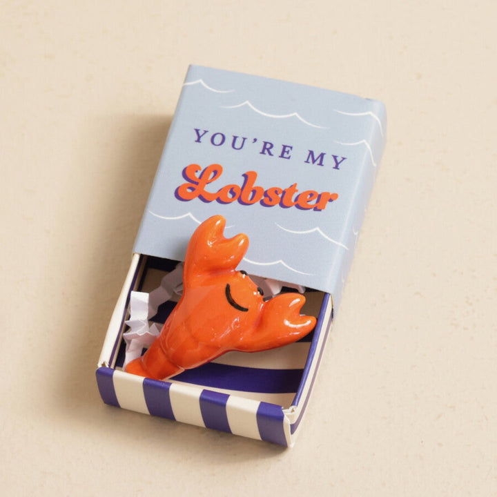 A small, red ceramic lobster figurine inside a blue matchbox-sized gift box. The box has the text 