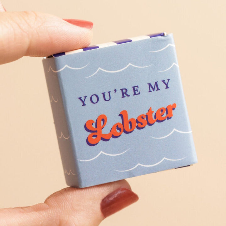 A small, red ceramic lobster figurine inside a blue matchbox-sized gift box. The box has the text 