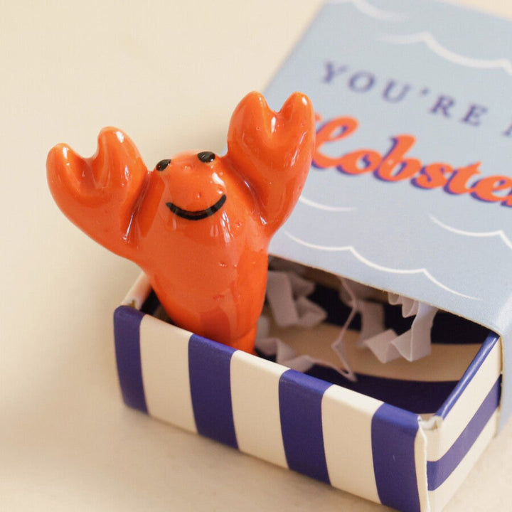 A small, red ceramic lobster figurine inside a blue matchbox-sized gift box. The box has the text 
