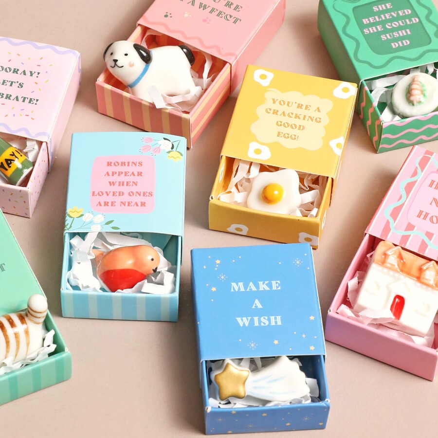 A collection of small matchbox-sized gift boxes with various ceramic figurines inside. Each box has a different message and design, such as "You're a Cracking Good Egg!" with a fried egg figurine, "Robins Appear When Loved Ones Are Near" with a robin figurine, and "Make a Wish" with a shooting star figurine.