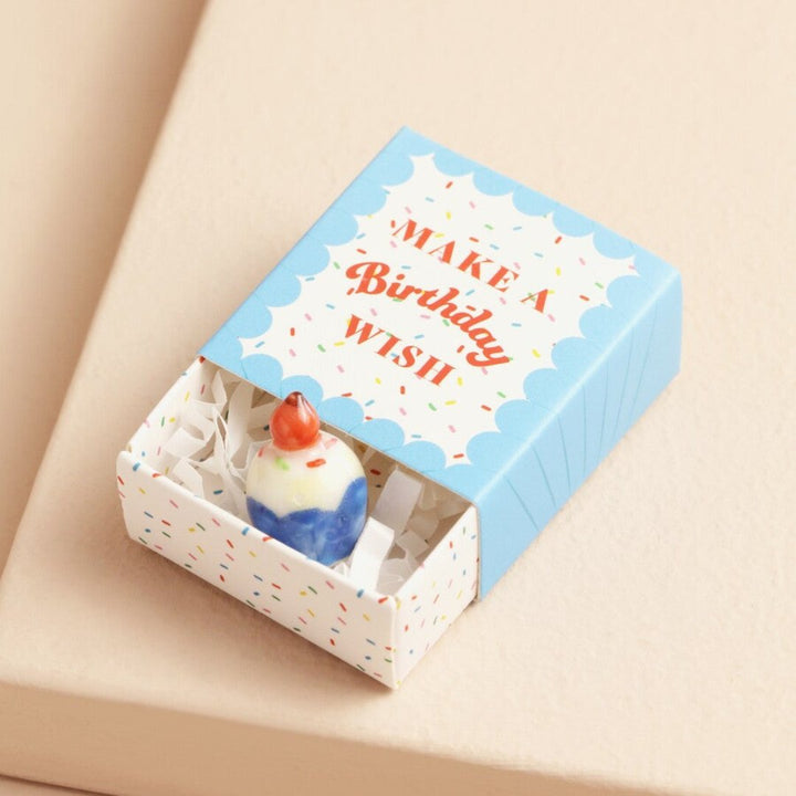 A small ceramic cupcake in a blue matchbox. The cupcake is frosted with white icing and has a red cherry on top. The matchbox has the text 