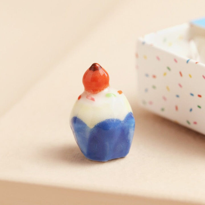 A small ceramic cupcake in a blue matchbox. The cupcake is frosted with white icing and has a red cherry on top. The matchbox has the text 